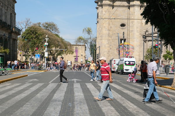 People crossing a sunlit street in Guadalajara's historic center, cathedral visible in the background