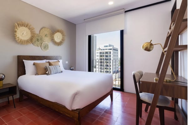 Studio bedroom with queen bed, woven wall art, wood nightstands, and open-plan kitchen and dining area visible through glass partition