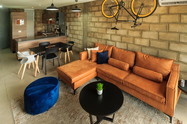 Sala de estar de loft moderno con sofá de cuero naranja, otomana de terciopelo azul, mesa redonda negra y dos bicicletas amarillas montadas en una pared de ladrillo