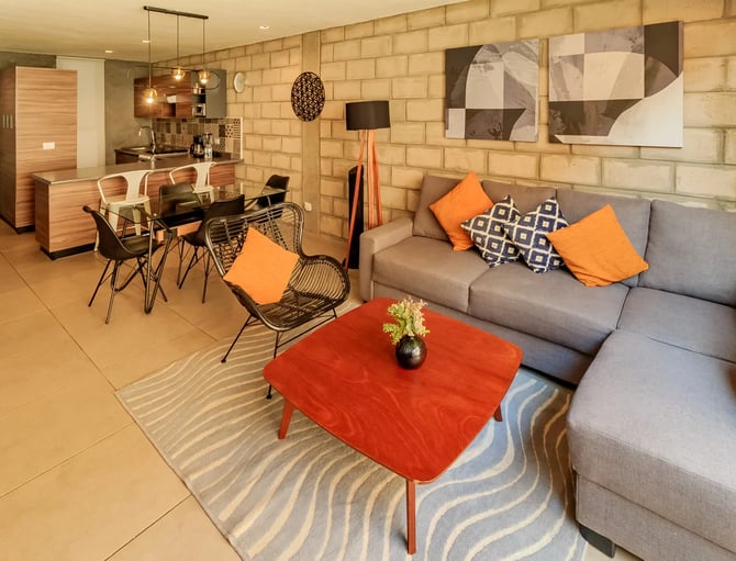 Open-plan living room with gray sofa, red coffee table, orange accents, and brick wall with dining area and kitchen visible in background